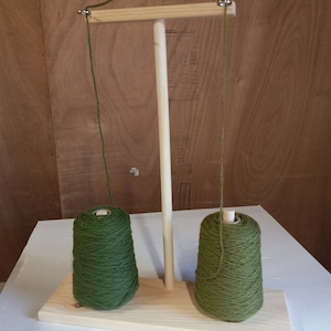May include: A wooden yarn holder with two spools of green yarn. The yarn holder has a wooden dowel and a crossbar with two hooks.