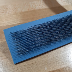 May include: A blue rectangular brush with a dense pattern of black bristles. The brush is designed for cleaning and grooming.