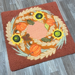 May include: A brown, orange, and green rug with a wreath design featuring pumpkins, sunflowers, and leaves. The wreath is on a brown background.