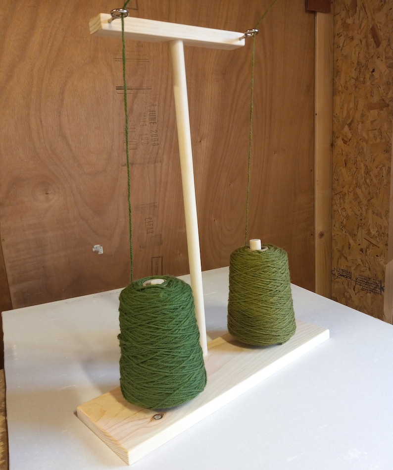May include: Wooden yarn holder with two spools of green yarn hanging from it. The yarn holder has a wooden base and a vertical post with a crossbar at the top.