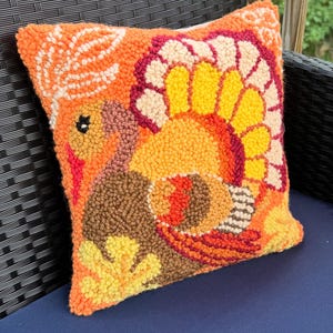 May include: A square, orange decorative pillow with a hand-tufted turkey design. The turkey features brown, yellow, orange, and red details. The pillow is placed on a blue surface and a black chair.