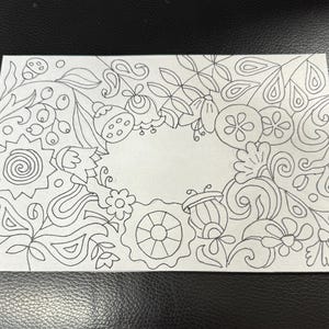 Peut inclure: Une carte rectangulaire blanche avec un dessin au trait noir de motifs floraux et abstraits. L'illustration présente des fleurs, des feuilles et d'autres éléments décoratifs entourant un espace rectangulaire vide au centre.