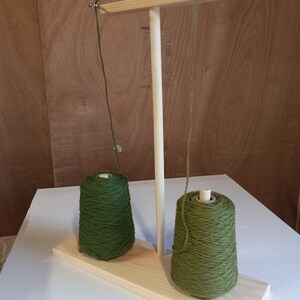 May include: A wooden yarn holder with two spools of green yarn. The yarn holder has a wooden base and a vertical wooden post with a crossbar at the top. The yarn is threaded through the crossbar and down to the spools.