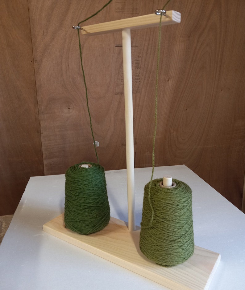 May include: A wooden yarn holder with two spools of green yarn. The yarn holder has a wooden base and a vertical wooden post with a crossbar at the top. The yarn is threaded through the crossbar and down to the spools.