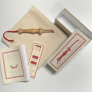 May include: A wooden punch needle, a red and white stitch gauge, a beige fabric, and a white booklet titled "The Oxford Punch Needle Handbook".