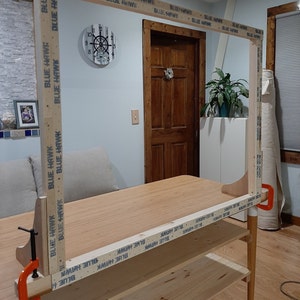 May include: A wooden frame is being assembled using clamps. The frame is made of wood planks and is being secured with blue tape that reads "BLUE HAWK". The frame is being assembled on a wooden table with a shelf.