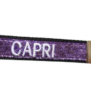 May include: A rectangular fabric keychain featuring a vibrant purple glitter surface with the name CAPRI embroidered in white capital letters. The strap is bordered by black fabric and attached to a silver-toned metal split ring via a metal clasp.