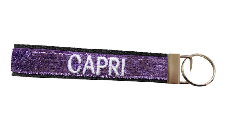 May include: A rectangular fabric keychain featuring a vibrant purple glitter surface with the name CAPRI embroidered in white capital letters. The strap is bordered by black fabric and attached to a silver-toned metal split ring via a metal clasp.