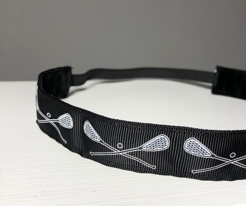 A black nonslip headband with white lacrosse sticks in an X formation.  The lacrosse stick pattern repeats along the length of the headband.  The back of the headband is lined with velvet and there is elastic that is worn toward the back of the head.