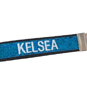 May include: A rectangular fabric keychain featuring a vibrant blue glitter surface with the name KELSEA embroidered in white capital letters. The strap is bordered by black fabric and attached to a silver-toned metal split ring hardware.