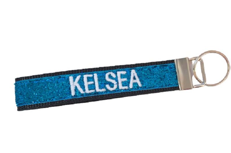 May include: A rectangular fabric keychain featuring a vibrant blue glitter surface with the name KELSEA embroidered in white capital letters. The strap is bordered by black fabric and attached to a silver-toned metal split ring hardware.