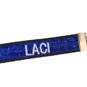May include: A rectangular fabric keychain featuring a vibrant blue glitter surface with the name LACI embroidered in white capital letters. The strap is bordered by black fabric and attached to a silver-toned metal hardware clasp and key ring.