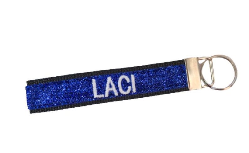 May include: A rectangular fabric keychain featuring a vibrant blue glitter surface with the name LACI embroidered in white capital letters. The strap is bordered by black fabric and attached to a silver-toned metal hardware clasp and key ring.