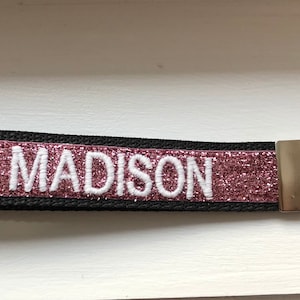 Black key fob with the name "MADISON" embroidered in white. The center band is a sparkling rose-colored glitter. A silver-colored metal key ring is attached to the end.