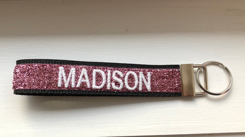 Black key fob with the name "MADISON" embroidered in white. The center band is a sparkling rose-colored glitter. A silver-colored metal key ring is attached to the end.