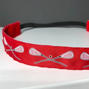 A red nonslip headband with white lacrosse sticks in an X formation.  The lacrosse stick pattern repeats along the length of the headband.  The back of the headband is lined with velvet and there is elastic that is worn toward the back of the head.