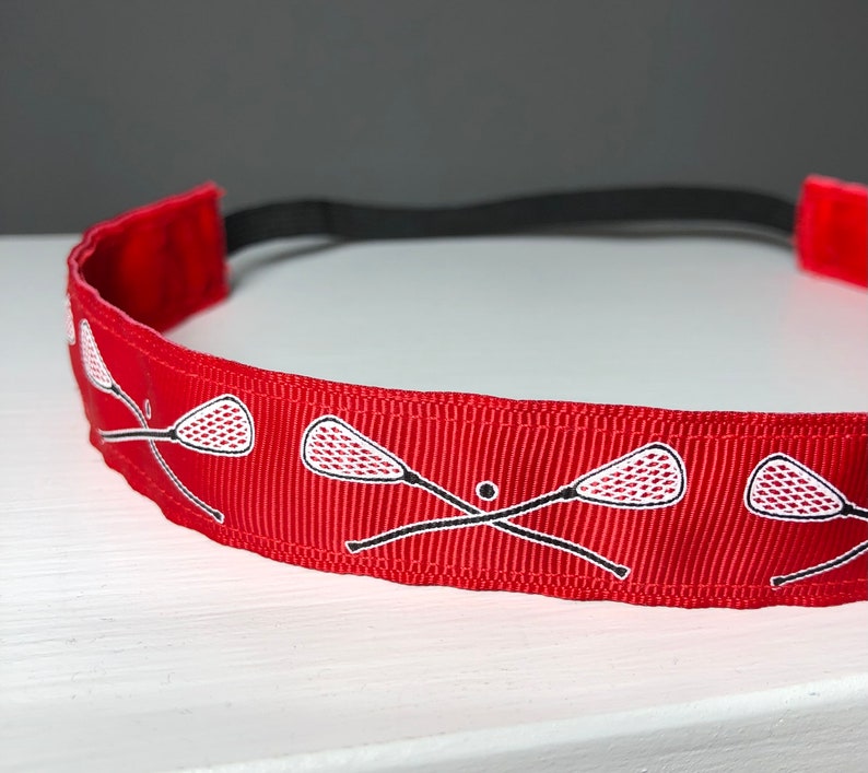 A red nonslip headband with white lacrosse sticks in an X formation.  The lacrosse stick pattern repeats along the length of the headband.  The back of the headband is lined with velvet and there is elastic that is worn toward the back of the head.