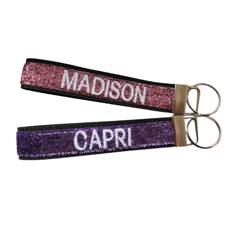 Two keychains with glitter straps. One is pink with the name "MADISON" embroidered in white, the other is purple with "CAPRI" in white. Both have black backing and silver colored key rings.