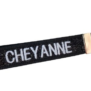 May include: A black glittery fabric keychain strap featuring the name CHEYANNE embroidered in white capital letters. The strap is secured with a silver-toned metal clamp and includes a matching circular metal split ring for attaching keys.