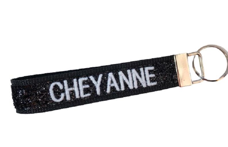 May include: A black glittery fabric keychain strap featuring the name CHEYANNE embroidered in white capital letters. The strap is secured with a silver-toned metal clamp and includes a matching circular metal split ring for attaching keys.