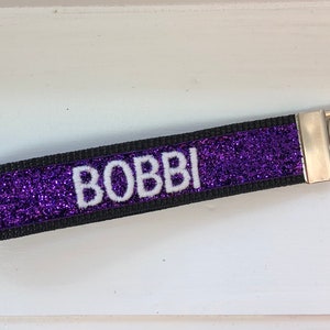A purple glitter keychain with the name "BOBBI" in white letters. The keychain has a black border and a silver metal key ring attachment. The design is simple and elegant.