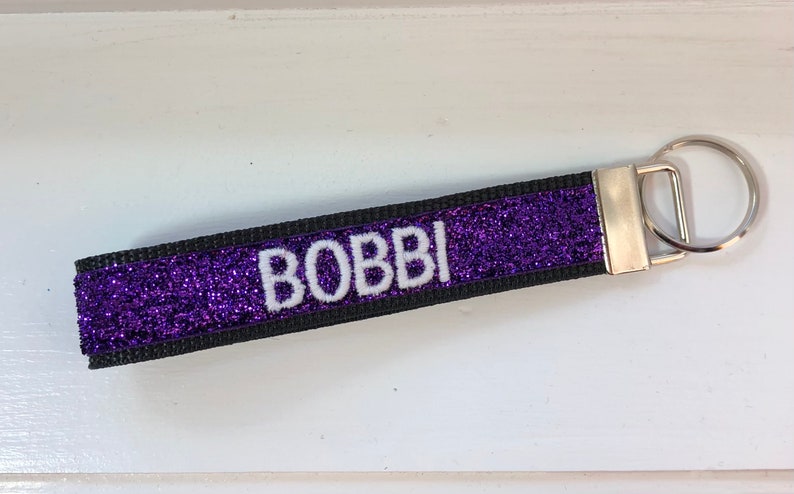 A purple glitter keychain with the name "BOBBI" in white letters. The keychain has a black border and a silver metal key ring attachment. The design is simple and elegant.