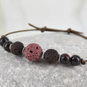 Puede incluir: Un brazalete de cuero marrón con cuentas de lava roja y marrón. El brazalete tiene una cuenta de acento plateada.