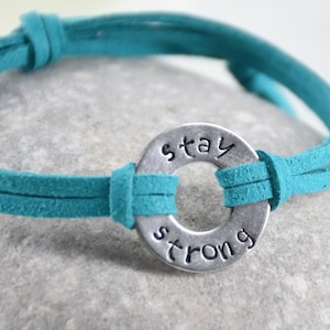Puede incluir: Pulsera de cuero gamuza turquesa con un dije de metal plateado que dice "stay strong".