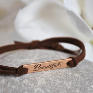 May include: A brown suede bracelet with a rose gold bar charm that says "Beautiful" in cursive.