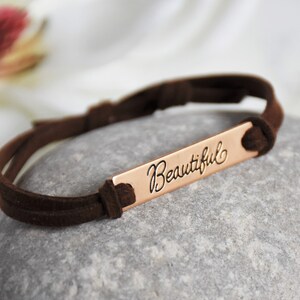 May include: A brown suede bracelet with a copper bar that says "Beautiful" in cursive.