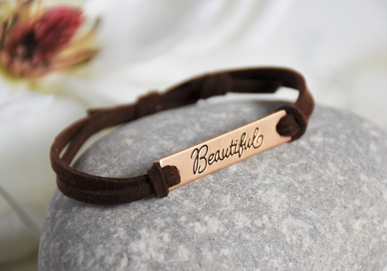 May include: A brown suede bracelet with a copper bar that says "Beautiful" in cursive.