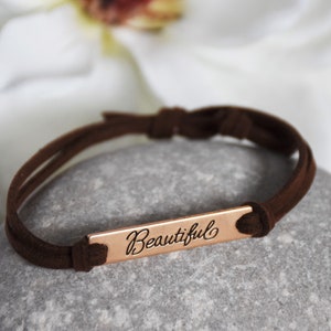 May include: A brown suede bracelet with a rose gold bar charm that says "Beautiful".