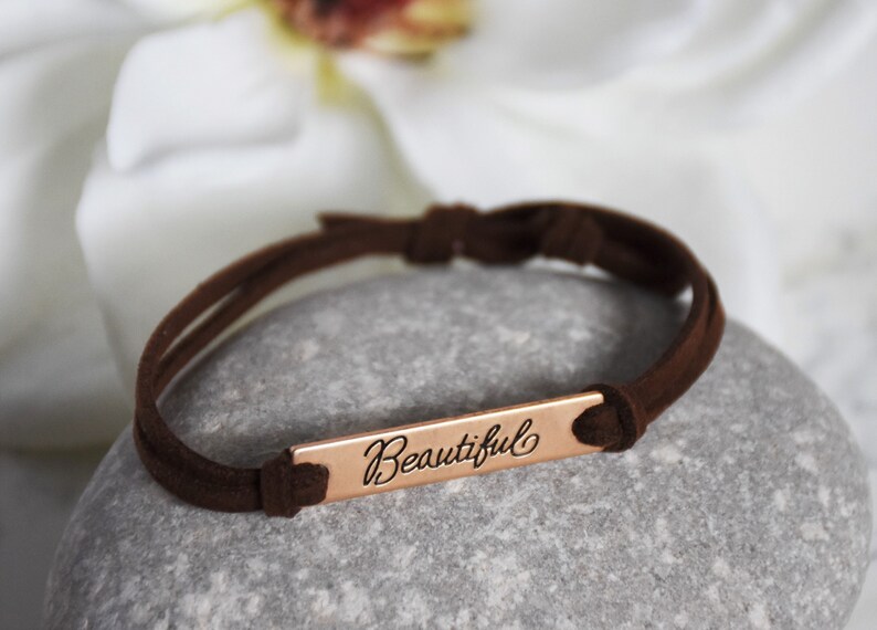 May include: A brown suede bracelet with a rose gold bar charm that says "Beautiful".