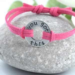 Può includere: Un braccialetto in pelle scamosciata rosa con un ciondolo d'argento che dice "you got this".