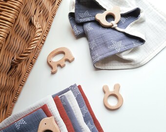 May include: A set of six muslin baby burp cloths in a variety of colors, including blue, white, and red. The cloths are folded and stacked on top of each other. There are also three wooden teethers in the shape of an elephant, a bunny, and a bird. The teethers are made of natural wood and have a smooth finish.