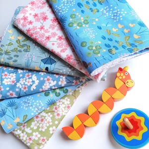 May include: A stack of colorful fabric squares with floral and water-themed patterns. A wooden caterpillar toy and a spinning top are also present. The fabrics feature designs of cherry blossoms, ducks, and water lilies. The toys are orange, yellow, and blue.