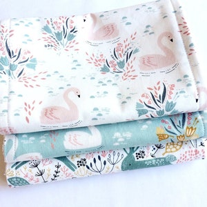 May include: Three fabric squares with a floral and swan pattern. The top square is white with pink swans and green floral designs. The middle square is light blue with white swans and yellow flowers. The bottom square is white with green and yellow floral designs and a pink turtle.