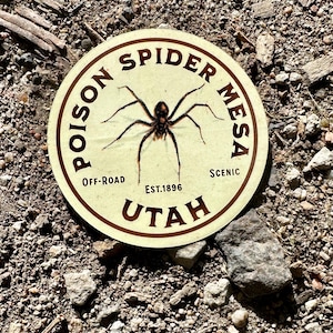 May include: A round sticker with a cream background and brown text. The sticker reads "POISON SPIDER MESA" around the top, with a spider graphic in the center. Below the spider are the words "OFF-ROAD" and "SCENIC". The word "UTAH" is at the bottom.