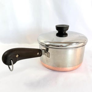 May include: A stainless steel saucepan with a copper bottom and a dark brown handle. The pot has a lid with a black knob. The handle has a metal loop for hanging.