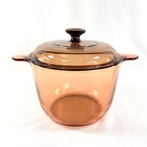 May include: A clear, amber-colored glass pot with a matching lid. The pot has two side handles and a rounded shape. The lid has a small knob handle. This cookware is suitable for cooking and serving.