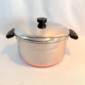 May include: A stainless steel cooking pot with a copper-colored base and a lid. The pot has two black handles and a black knob on the lid. The pot is designed for cooking.