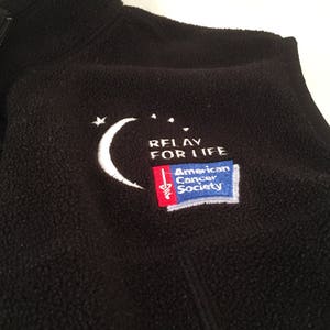 May include: Black fleece vest with a high collar and a partial zipper. The vest features an embroidered design with a white crescent moon, stars, and the text "RELAY FOR LIFE" and "American Cancer Society" in a rectangular patch.