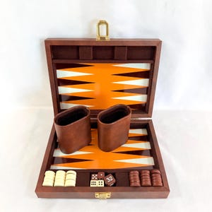 May include: Open wooden backgammon set with a brown exterior and orange and white playing surface. Includes brown leather dice cups, dice, and game pieces. The set is displayed on a white background.
