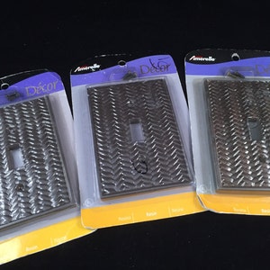 May include: Three decorative light switch covers in a package. The covers have a textured, woven design and are in shades of silver, bronze, and brown. The packaging includes the word "Décor" and the brand name "Amerelle".
