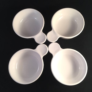 May include: Four white ceramic serving bowls with a handle-like extension. The bowls are arranged in a square pattern against a black background. The bowls are round with a small, rounded handle.
