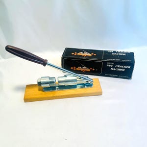 May include: A silver nutcracker machine with a brown wooden handle and a wooden base. The machine is in its original box, which is black with white text that reads "Deluxe Nut Cracker Machine, Data No 23557, Made in Taiwan."