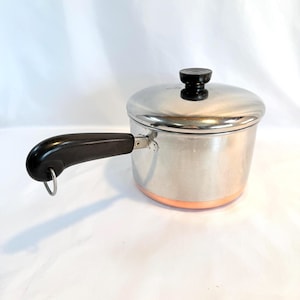 May include: A stainless steel saucepan with a lid and a black handle. The pot has a copper-colored base and a black knob on the lid. The handle has a loop at the end. Kitchenware.