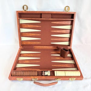 May include: A brown backgammon set in a briefcase-style case with a felt interior. The board features alternating brown and cream-colored triangles. Included are brown and cream checkers, a doubling cube, two dice, and a brown leather dice cup.