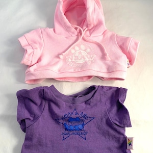 May include: Two doll-sized tops: a light pink hooded sweatshirt with "NAAW" and paw print graphic, and a purple t-shirt with a blue graphic of Noah's Ark and the text "NOAH'S ARK ANIMAL WORKSHOP".