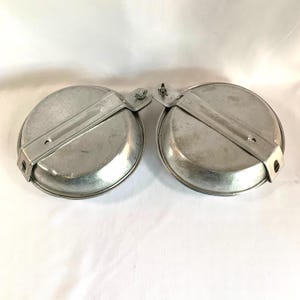 May include: Two silver aluminum camping mess kits with a hinged handle. The kits are made of metal and have a round shape.